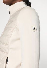 Ljust beige jacka med en figursydd design, som har en vadderad frampanel, släta ärmar och en cirkulär logotyp på ärmen.