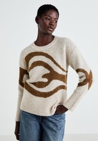 Beige genser med brun abstrakt design, ribbestrikkede mansjetter og hem, laget av et mykt materiale. Kombinert med lyseblå denimjeans.