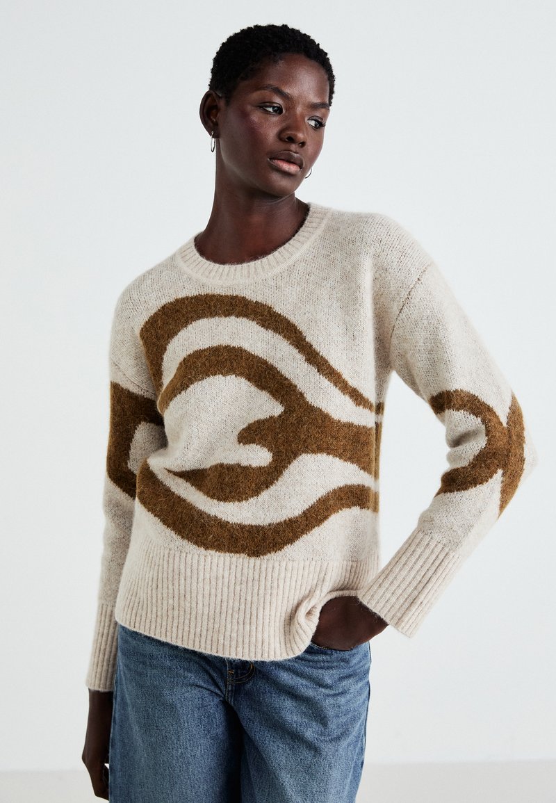 Beige genser med brun abstrakt design, ribbestrikkede mansjetter og hem, laget av et mykt materiale. Kombinert med lyseblå denimjeans.