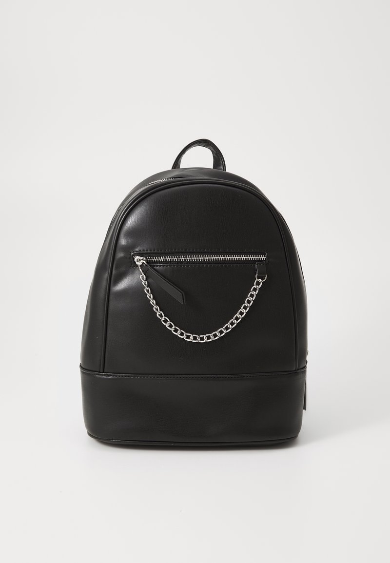 Sac à dos compact en cuir noir avec poche zippée à l'avant et chaîne argentée décorative sur un fond clair uni.