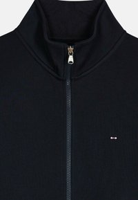 Veste zippée noire avec col haut, texture côtelée et fermeture éclair argentée. Présente un petit logo rose sur la zone gauche de la poitrine.