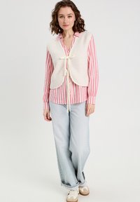 Gilet en polaire blanc avec un fermoir à nœud papillon, porté par-dessus une chemise rayée rose et blanche. Un jean large bleu clair et des baskets beiges complètent le look.