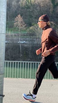 Δρομέας με καφέ μπουφάν, μαύρο παντελόνι και σκούφο τρέχει κατά μήκος μονοπατιού δίπλα στον ποταμό, πλαισιωμένου από μεταλλικό κιγκλίδωμα και δέντρα.