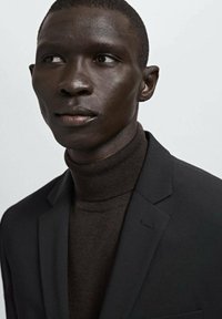 Donkere man met een zwarte blazer en een donkerbruine coltrui, die iets naar links kijkt tegen een effen lichte achtergrond.