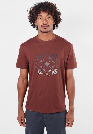Homme aux cheveux bouclés portant un T-shirt marron avec un motif floral et un texte indiquant "Surf Gear est. 1985" et un short foncé.