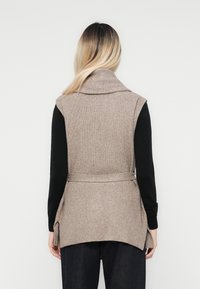 Beige geribbelde mouwloze vest met een brede kraag, ceintuur in de taille, voorzien van verstelbare zijkanten en een gestructureerde afwerking, gedragen over een zwart langemouwen shirt.