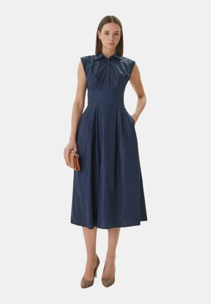 Donna in piedi che indossa un abito midi senza maniche blu navy con colletto, che tiene una piccola pochette marrone e indossa tacchi alti color cuoio.
