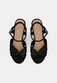 Sandales noires avec plusieurs lanières découpées, semelle plate et boucle de cheville ajustable. Texture lisse avec un semelle intérieure beige et un détail de logo.