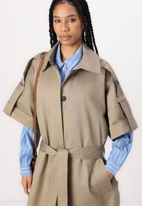 Khaki trench coat so štruktúrovaným dizajnom, krátkymi rukávmi a pásikom v páse. Skombinovaný s modrou pruhovanou košeľou pod ním.