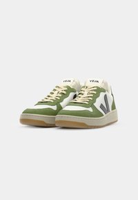 Grønne og hvite joggesko laget av semsket skinn og lær, med beige lisser og såle i gummigummi. "VEJA"-logo på tungen og siden.