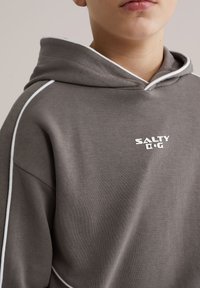 Hanorac gri din amestec de bumbac, cu cordon alb. Are un logo pe față, glugă mare și croială lejeră. Potrivit pentru ținute casual.