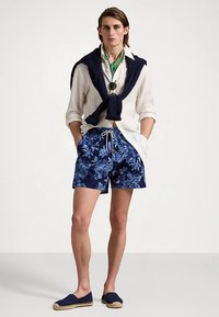 Νεαρός άνδρας στέκεται με τα χέρια στις τσέπες, φορώντας λουλουδάτα σορτς για κολύμπι, λευκό πουκάμισο, ναυτικό πουλόβερ ριγμένο στους ώμους και εσπαντρίγιες.