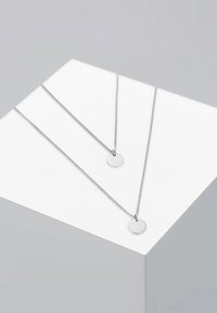 To sølv halskæder med cirkulære vedhæng, der har et strømlinet, minimalistisk design. Kæderne er sarte med en glat, poleret finish.