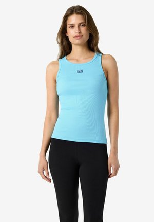 Femme portant un haut sans manches bleu clair et un pantalon noir sur un fond blanc uni, regardant vers l'avant avec une expression neutre.