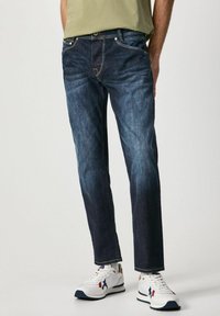 Jeans de mezclilla azul oscuro con corte recto, que cuentan con cinco bolsillos, costuras en contraste y una cintura con trabillas, combinados con zapatillas blancas.