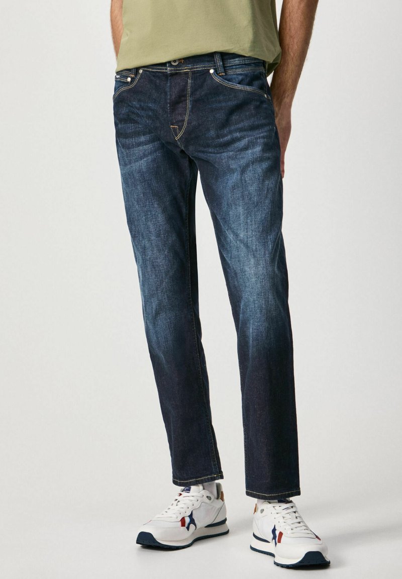 Jeans de mezclilla azul oscuro con corte recto, que cuentan con cinco bolsillos, costuras en contraste y una cintura con trabillas, combinados con zapatillas blancas.
