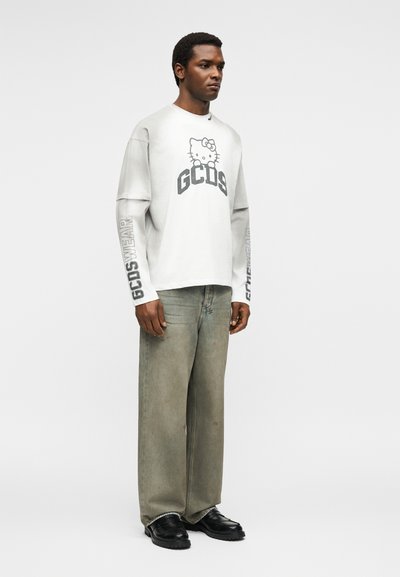 Άνδρας που φοράει λευκό πουκάμισο μακρυμάνικο με το λογότυπο Hello Kitty και GCDS, ξεβαμμένο φαρδύ τζιν και μαύρα παπούτσια, στέκεται μπροστά σε απλό φόντο.