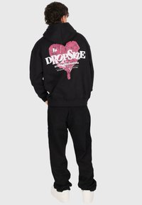 Person wearing black hoodie with pink heart and white "DROPSIZE" text on back, black pants, and white sneakers, standing against plain background.