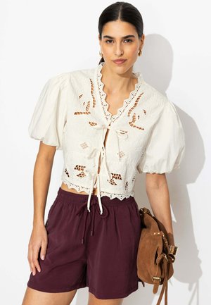 Femme portant un chemisier blanc en dentelle œillet noué devant avec des manches bouffantes, un short bordeaux à cordon et tenant un sac à dos en daim marron.