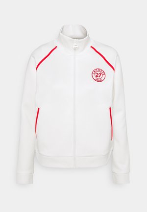 Veste blanche à fermeture éclair avec passepoil rouge sur les épaules et les poches, et logo circulaire rouge "Lacoste 27 Depuis" sur la poitrine gauche.