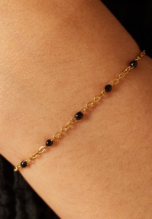 Gouden kettingarmband met kleine zwarte kralen, gedragen om een lichtbruine pols. Gladde textuur met afwisselende schakels en kralen.
