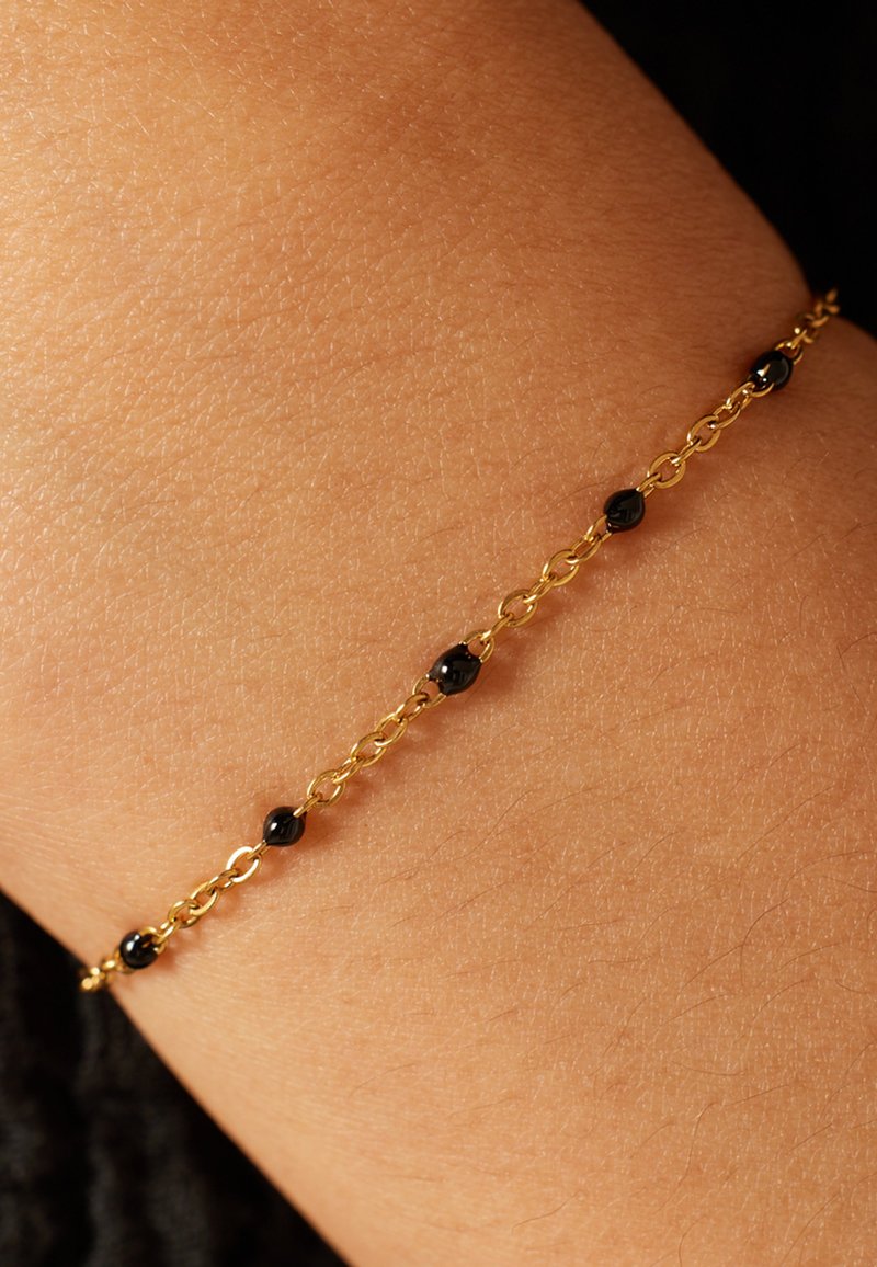 Gouden kettingarmband met kleine zwarte kralen, gedragen om een lichtbruine pols. Gladde textuur met afwisselende schakels en kralen.