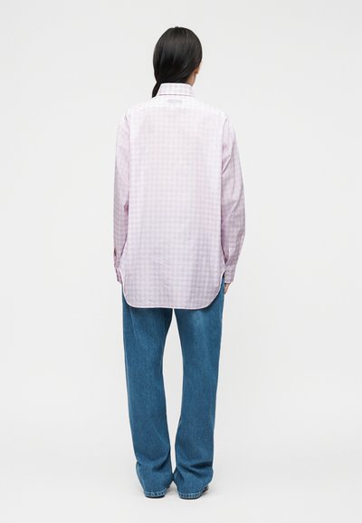 Γυναίκα με μακριά σκούρα μαλλιά σε χαμηλή αλογοουρά, που φοράει ροζ ανοιχτό ροζ καρό πουκάμισο μακρυμάνικο και μπλε τζιν φαρδιάς γραμμής, στεκόμενη με την πλάτη προς τα μας.