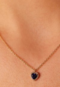 Collarín de tono de piel adornado con un collar de cadena de oro que cuelga de un colgante en forma de corazón con una gema azul, rodeado de pequeñas piedras transparentes.