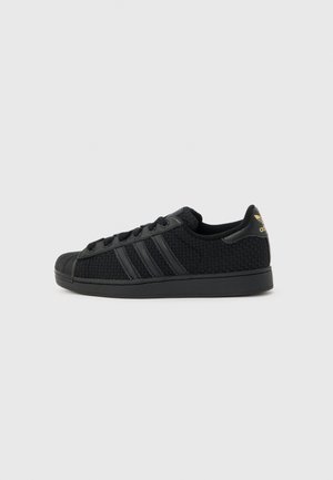 Pantof sport Adidas negru cu textură țesută, vârf de cauciuc negru, șireturi negre și logo auriu trefoil pe eticheta de pe călcâi, văzut din partea exterioară.