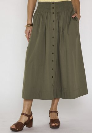 Femme portant une jupe midi vert olive à boutons sur le devant avec une taille smockée, associée à des sandales marron à talons et des bracelets dorés.