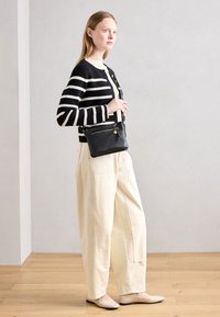 Cardigan a righe bianche e nere, camicia crema, pantaloni larghi color avorio e scarpe chiare. La borsa è in pelle nera con dettaglio a zip.