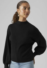 Maglione nero a maglia con collo alto e maniche a palloncino, caratterizzato da una finitura testurizzata. Abbinato a jeans blu chiaro per creare contrasto.