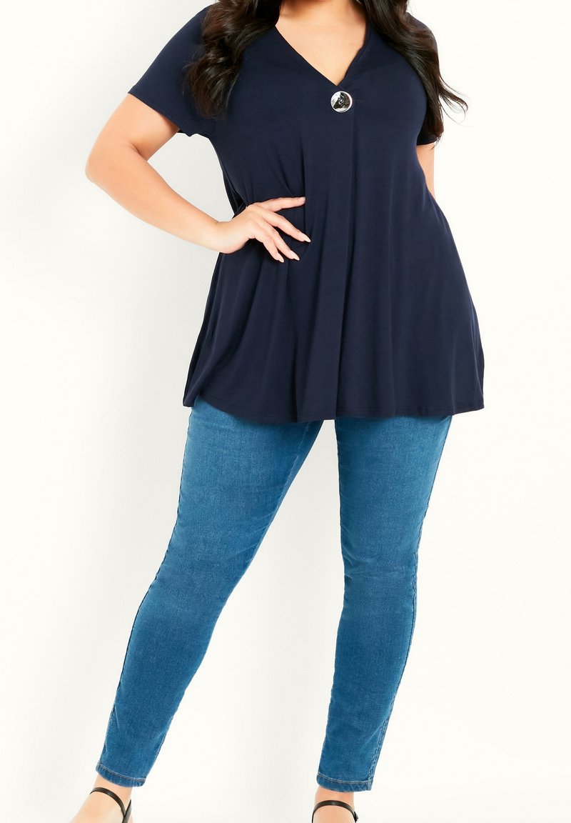 Femme portant un haut bleu marine à manches courtes avec un grand bouton et un jean skinny bleu clair, posant avec une main sur la hanche.