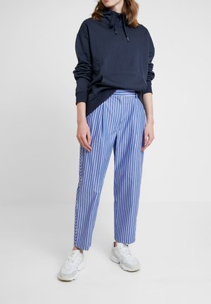 Personne portant un sweat à capuche bleu marine, un pantalon à rayures verticales bleues et blanches, et des baskets blanches, debout devant un fond clair uni.