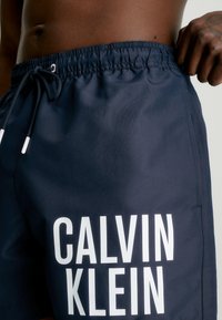 Pantaloni da bagno navy con cinturino elastico, chiusura con lacci e logo bianco "CALVIN KLEIN" in evidenza sul lato anteriore sinistro. Texture liscia.