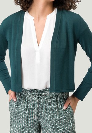 Femme portant une blouse blanche à col en V, un cardigan ouvert vert foncé et un pantalon taille haute à motifs, les mains dans les poches.