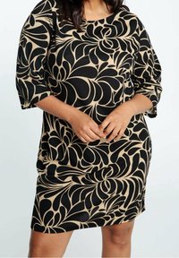 Femme portant une robe midi à motifs noirs et beiges avec des manches trois-quarts, les mains doucement jointes devant.