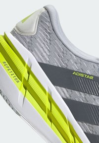 Gros plan d'une chaussure de course grise et jaune fluo avec une tige en tissu texturé et une semelle blanche, étiquetée "Repetitor" et "Adistar" sur le côté.