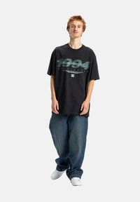 Čierne oversize tričko s grafickým textom a logom na prednej strane. Tmavomodré voľné džíny. Model má na sebe biele tenisky.