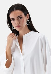 Weißes Hemd mit durchgehender Knopfleiste, Stehkragen, langen Ärmeln und lässiger Passform. Das Model trägt übergroße silberne Ohrringe mit wellenförmigem Design.