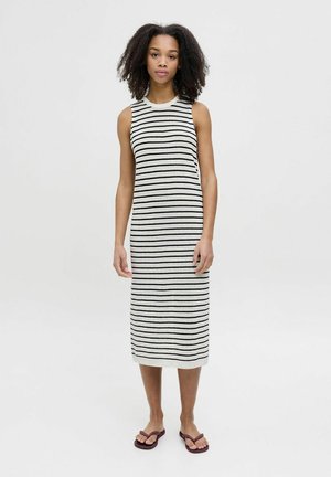 Vestido sin mangas a rayas en blanco y negro, de longitud hasta la rodilla y con una silueta ajustada. Combinado con chanclas moradas. Textura de punto suave.