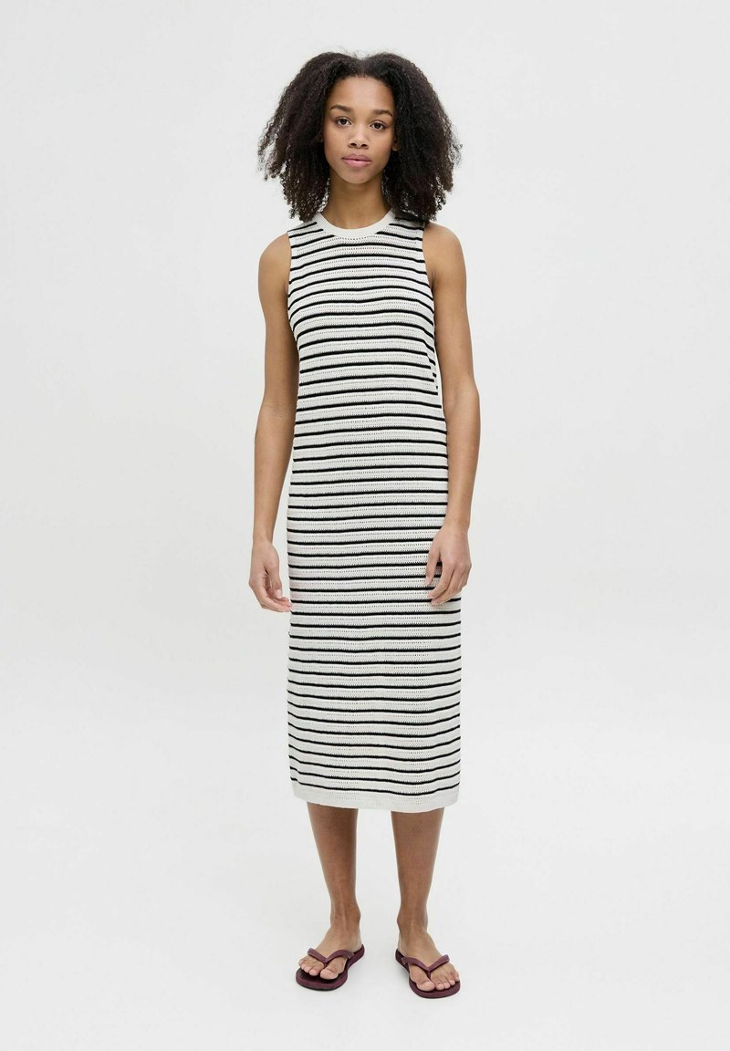 Vestido sin mangas a rayas en blanco y negro, de longitud hasta la rodilla y con una silueta ajustada. Combinado con chanclas moradas. Textura de punto suave.
