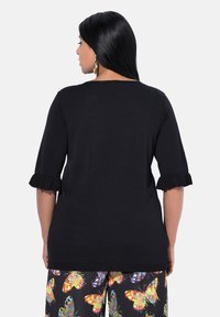 Schwarzes Strickoberteil mit kurzen, rüschengesäumten Ärmeln und rundem Ausschnitt. Im Hintergrund sind bunte Hosen mit Schmetterlingsmuster zu sehen.
