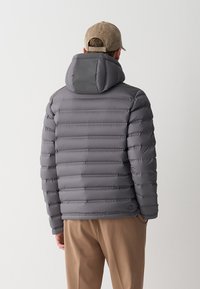 Uomo che indossa una giacca imbottita grigia con cappuccio e un berretto beige, in piedi con le mani in tasca su uno sfondo bianco semplice.