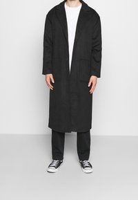 Persona che indossa un lungo soprabito nero sopra una camicia bianca e pantaloni neri, in piedi con le mani rilassate lungo i fianchi e sneakers nere con lacci bianchi.