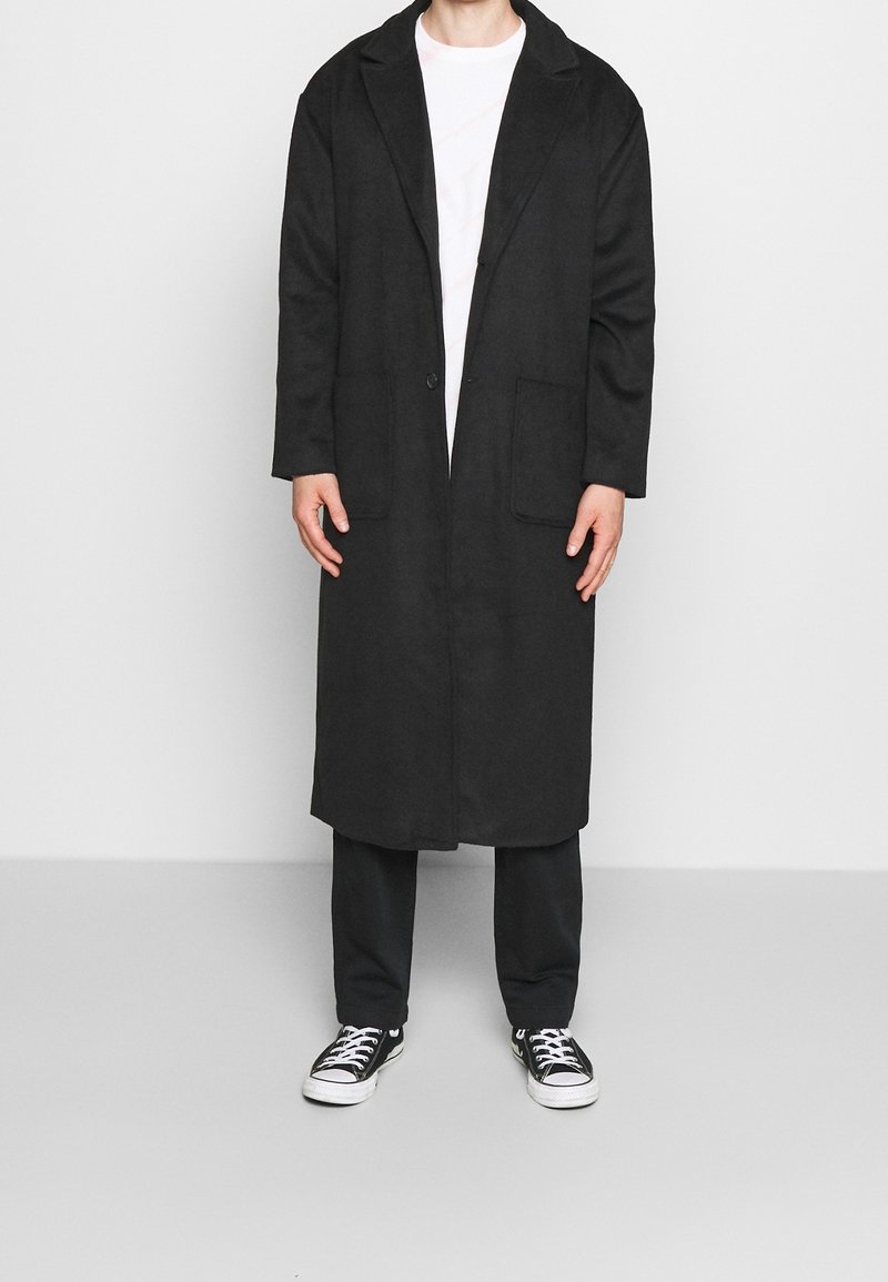 Persona che indossa un lungo soprabito nero sopra una camicia bianca e pantaloni neri, in piedi con le mani rilassate lungo i fianchi e sneakers nere con lacci bianchi.