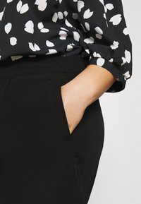 Czarne spodnie z elastycznym pasem i kieszeniami po bokach, w połączeniu z czarną bluzką ozdobioną białymi liśćmi i marszczonymi rękawami.
