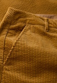 Pantalon en velours côtelé jaune moutarde avec une texture à côtes verticales, une poche avant et des détails de couture visibles. Doublure intérieure lisse.