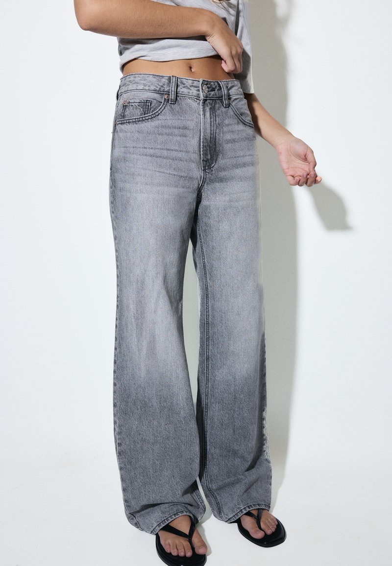 Personne porte un jean gris délavé taille haute à jambes larges et des tongs noires, avec un haut gris clair court montrant le ventre découvert.