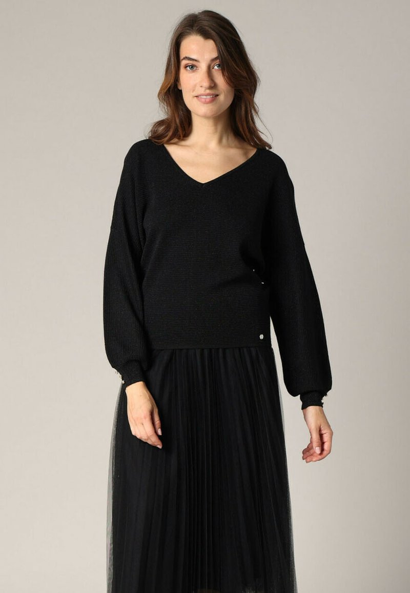 Pull en maille noire avec un col en V, des manches ballon et une texture côtelée, associé à une jupe en tulle plissé noire, ornée de boutons en métal argenté.